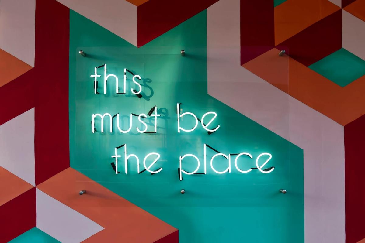 A sign with neon letters saying “this must be the place” on a colourful background, the text referring to companies offering an open vacation policy