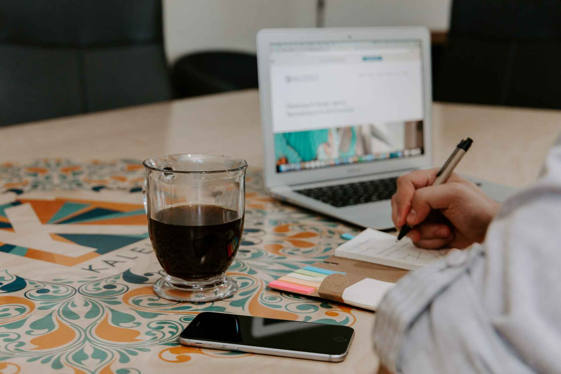 person sitting at a desk with a laptop, coffee, and notepad while writing self feedback