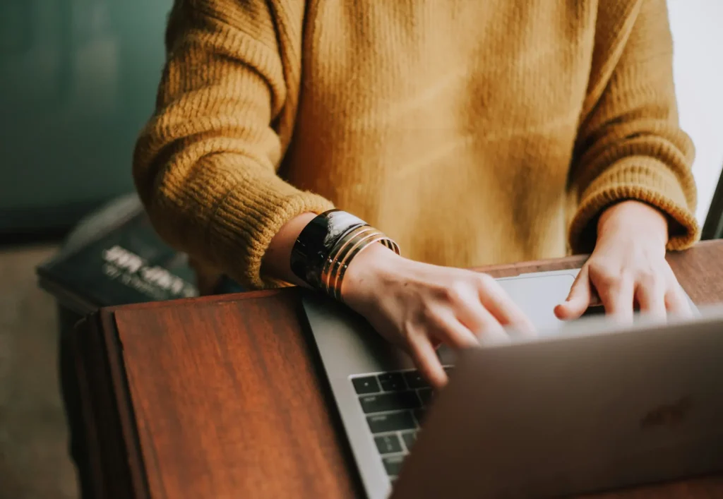 Nahaufnahme einer Person in einem gemütlichen Pullover, die an einem Holztisch auf einem Laptop tippt – ein Sinnbild für Remote-Arbeit und einen skalierbaren Einstellungsprozess.